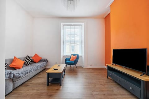 Living room with a grey sofa dressed with orange cushions, a blue armchair, a central coffee table, a large TV on a dark media unit, and a bold orange feature wall opposite the window.