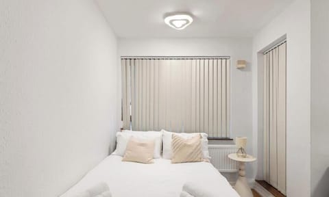 This compact, minimalist bedroom features a white-clad bed tucked snugly between bright walls under a triangular ceiling light. Vertical blinds and a pedestal side table complete the room’s clean, modern aesthetic.