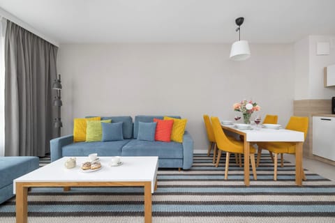A cozy seating area features a blue sofa adorned with colorful pillows, paired with a wooden coffee table. In the background, a dining set with vibrant yellow chairs sits adjacent to a modern kitchenette.