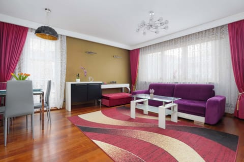 A bright living room with a purple sectional sofa and a white coffee table. Large windows with red curtains let in plenty of natural light.