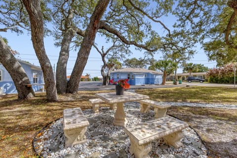 Outdoor seating area with a stone table and benches set beneath leafy trees beside a blue house.