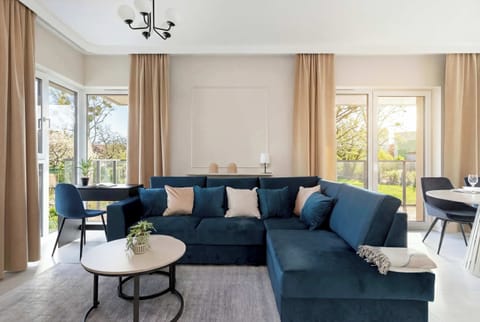 A spacious living room features a large, navy blue corner sofa adorned with soft pillows, creating a perfect spot for relaxation. Large windows let in plenty of natural light, highlighting the modern interior and light-colored flooring.