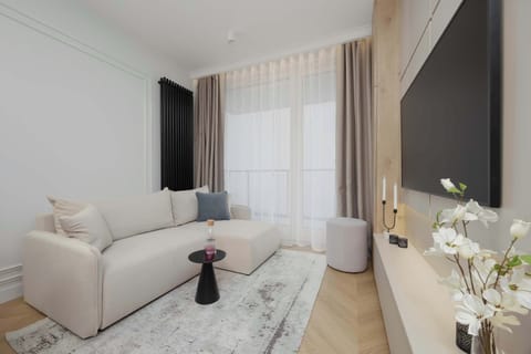 A bright living room featuring a cream-colored sectional sofa, a textured grey rug, and a large window with floor-to-ceiling beige curtains.