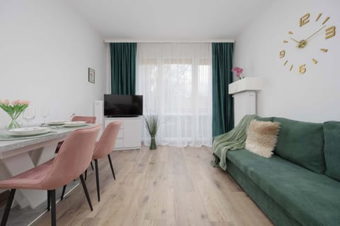 A bright living and dining area with light wooden floors and white walls. A green sofa stands against the wall near a small dining table with pink upholstered chairs. Large windows with dark green curtains let in natural light.