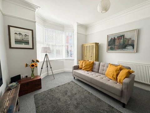 Grey sofa with mustard cushions in the bay window corner of the sitting room, with a yellow louvred-door cabinet, floor lamp, and two framed paintings on the walls.