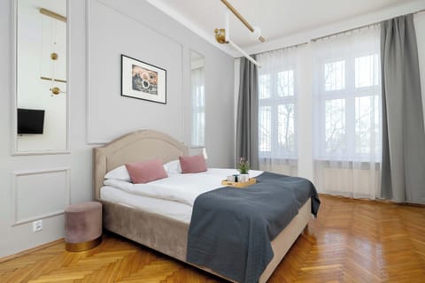 A bright and elegant bedroom featuring a large bed with a grey throw and pink decorative pillows. The room is decorated with classic wall moldings, a golden pendant light, and warm herringbone parquet flooring.