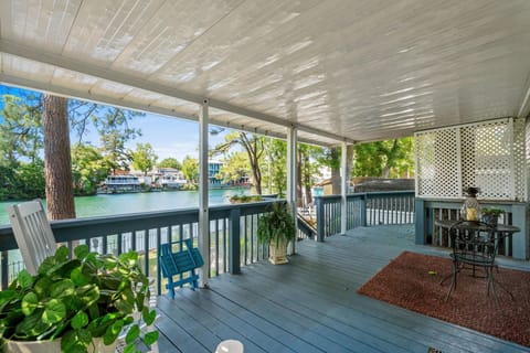 Covered porch with gray wood floorboards, blue railing, black metal bistro set, and lake views beyond trees.