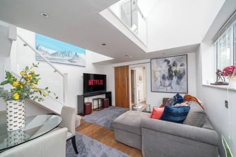Bright and modern living room with a skylight, plush sectional sofa, and colorful accent pillows. Features include a wall-mounted TV, contemporary artwork, and a glass dining table. Stylish and cozy with plenty of natural light and open space.