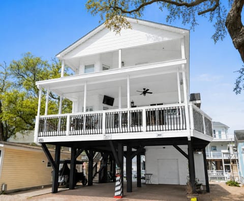 Two-story elevated white house with wraparound balcony, black metal railings, ceiling fan, and outdoor TV.