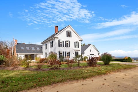 A classic white New England farmhouse set against open fields — your home base for exploring the Lakes Region.