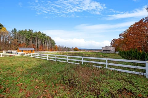 White post-and-rail fencing borders the open fields, with fall foliage and a glimpse of the mountains visible in the distance.