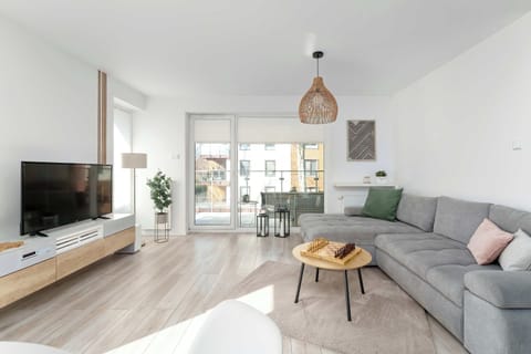 A bright, modern living room featuring a large grey corner sofa, a wooden coffee table, and a wall-mounted TV. Large glass doors lead out to a sunlit balcony, filling the space with natural light.