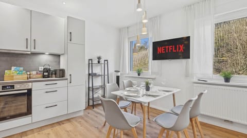 Weißer Esstisch mit vier grauen Stühlen, Fernseher mit Netflix-Logo, offene Küche im Hintergrund.