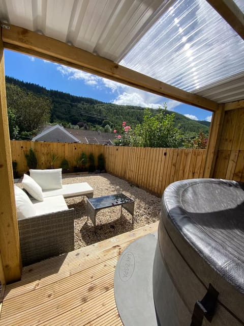 Rear garden with a timber pergola, outdoor sofa set, gravel area, decked hot tub platform, and a tree-covered hillside beyond the fence.