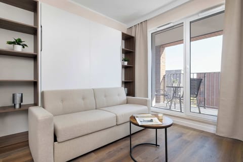 A bright living room features a light-colored sofa, a round coffee table, and large glass balcony doors. Soft curtains and neutral tones create a calm and welcoming atmosphere.