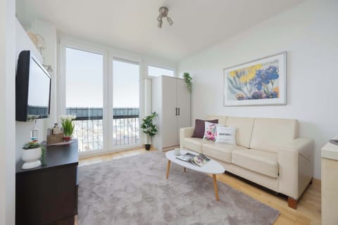 A bright living room features a cream-colored sofa and a dark media console under a wall-mounted television. A large balcony door lets in plenty of natural light, illuminating the grey rug and colorful wall art.