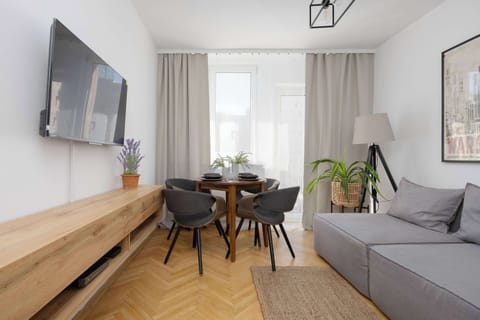 The cozy living area combines the functions of a dining room and a living room, featuring a gray corner sofa and a round table with four chairs. A large TV is mounted above the wooden TV stand.