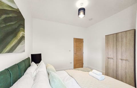 A modern bedroom featuring a queen bed with an emerald quilted headboard, grey patterned accents, and stacked-ring lamps. An abstract landscape hangs above the bed, flanked by white-shuttered windows and grey curtains.