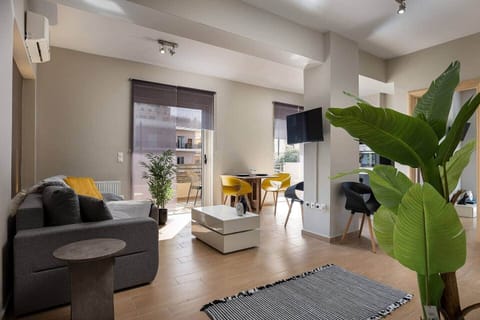 Bright open-plan living area with a comfortable sofa, wall-mounted TV, and a compact dining table. Large balcony doors bring in natural light and connect the room to an outdoor seating area, adding an easy indoor-outdoor feel.