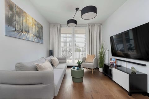 A bright living room with a comfortable sofa, a TV mounted on the wall, and a small coffee table. Natural light comes through large windows, creating a cozy and inviting atmosphere.