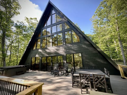 Spacious wooden deck with black A-frame cabin exterior, outdoor dining table, rocking chairs, and hot tub surrounded by trees.