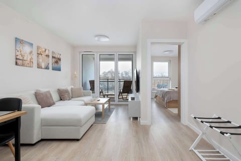 A bright living room connected to a kitchenette, featuring a white corner sofa and modern, light-colored floor panels