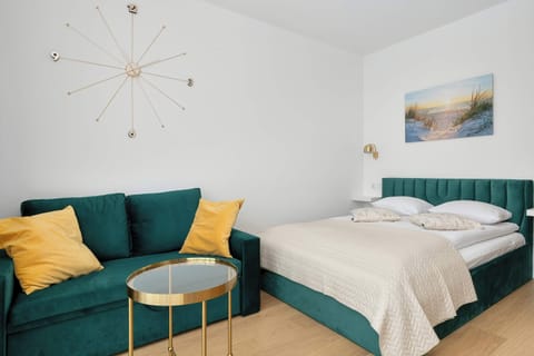 A stylish studio apartment combines a cozy sleeping area with a vibrant teal velvet sofa. Decorative gold accents and a minimalist wall clock add a touch of sophistication to the bright, sunlit room.