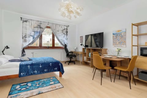 A bright studio space with a double bed, a small desk, and a dining area near the window. Large windows fill the open layout with natural light.