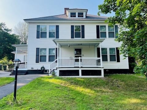 A charming white colonial home with classic green shutters sits on a peaceful street, offering a welcoming front porch.