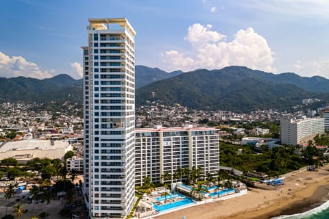 Elevated drone shot of the sleek, modern white residential skyscraper standing prominently along the sandy shoreline.