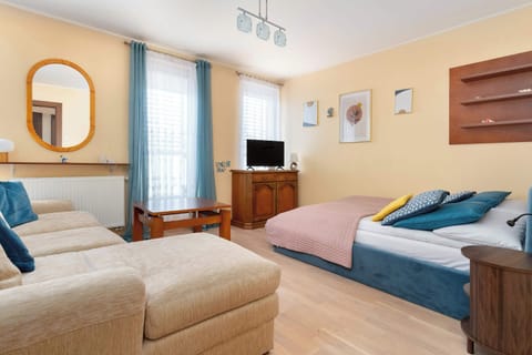 A bright living room features a large, comfortable L-shaped beige sofa and a wooden coffee table. In the background, a single bed with blue and yellow accents is visible, suggesting a multifunctional studio space.