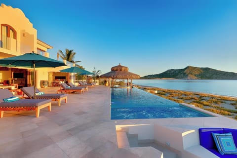 Stunning beachfront villa with infinity pool overlooking pristine waters and distant mountains in golden evening light.