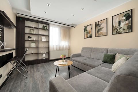 A spacious living area features a large grey L-shaped sofa, a minimalist wooden coffee table, and walls adorned with framed botanical prints.