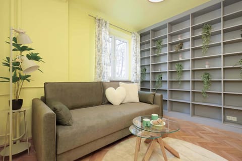 A cozy living room featuring a plush sofa, a wooden coffee table, and an expansive floor-to-ceiling bookshelf filled with diverse books and integrated plants, set against a pale yellow wall.