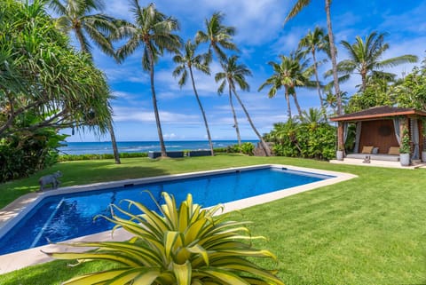 A stunning oceanfront property featuring a sparkling pool, lush lawn, and swaying palms. The pavilion and manicured grounds frame breathtaking views of the Pacific.