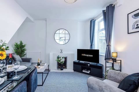 A bright, modern living room with gray sofa and armchair, glass coffee table, and a cozy fireplace topped with a round mirror. Natural light flows in through large windows, while a dining table with sunflowers adds warmth and charm.
