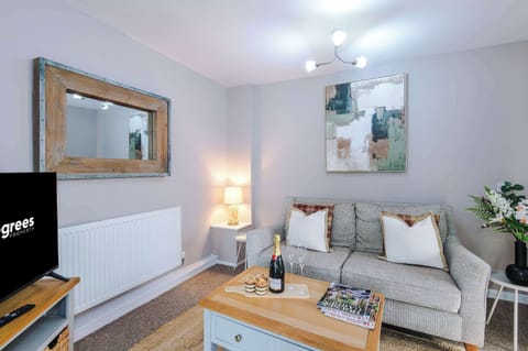 A cozy modern living room with a gray sofa, patterned throw, and champagne on the coffee table. Abstract art and a wooden-framed mirror add style, while soft lighting creates a warm, inviting atmosphere.