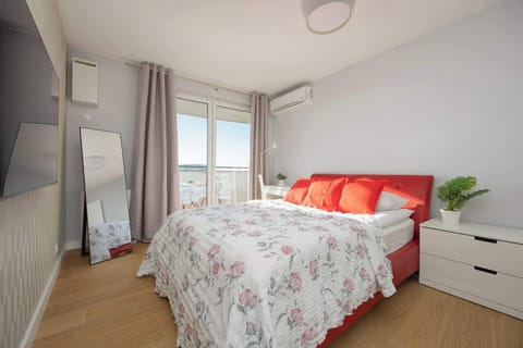 A vibrant primary bedroom features a large bed with a floral duvet and bold red pillows. A tall standing mirror and white bedside tables complete the bright, airy space.