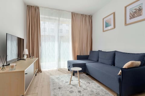 A bright living room with a blue sofa, a small round coffee table, and a TV on a wooden stand near a large window with curtains.