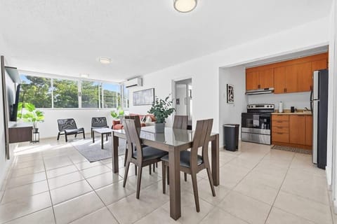 Spacious open-concept dining and living area with a glass dining table, large windows with greenery views, cozy seating area, and a nearby kitchen with warm wood cabinets and stainless appliances.