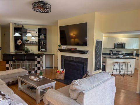 Light wood floors connect the living room with beige sofas, a black-tiled fireplace, and a wall-mounted TV.