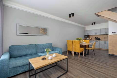 A bright open-plan living room features a comfortable teal blue sofa, a wooden coffee table, and a dining area with vibrant yellow chairs.