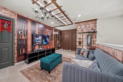 Welcome to a cozy retreat with rich wallpaper that evokes a classic library. Enjoy the plush teal ottoman and soft gray sofa, all enhanced by a stylish chandelier that brings a touch of sophistication to your relaxation space.