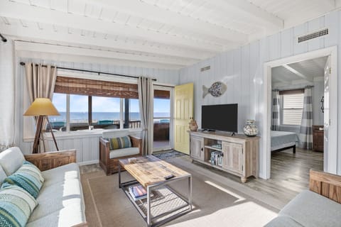 Bright coastal living room with sofas, chairs, TV, coffee table, floor lamp, wood ceiling beams, and large windows plus a door opening to a deck with ocean views.