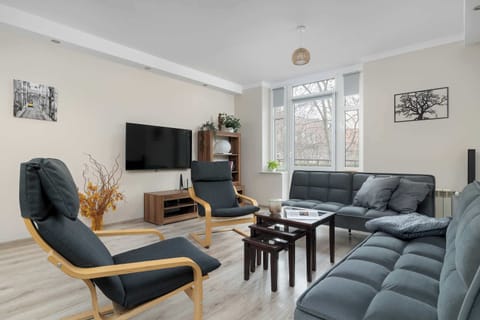 A spacious living room featuring a comfortable grey L-shaped sofa and two stylish wooden armchairs. A large window fills the space with natural light, highlighting the modern decor.