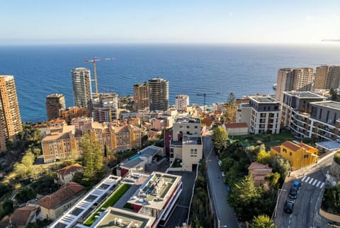 An elevated view across a dense urban roofscape of mixed-height buildings leading down to a broad stretch of sea under a clear blue sky.