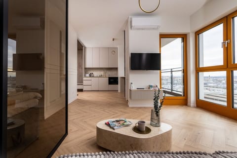 A spacious living room connected to a kitchenette, featuring a stylish stone coffee table as its centerpiece. Large windows and access to the balcony allow plenty of natural light to flood the interior.