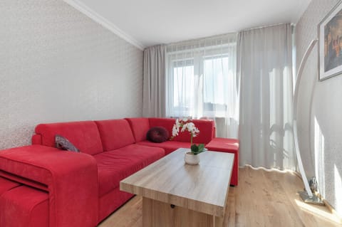 A vibrant red corner sofa serves as the focal point of this bright living room, complemented by a light wood coffee table. The space is filled with natural light from a nearby window draped in sheer white curtains.