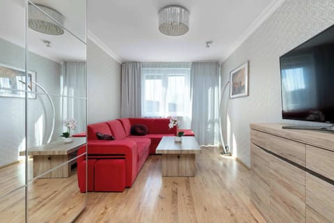 This wide-angle shot of the living room highlights a long, light-wood dresser and a large television. The red sectional is visible in the background, showing the flow of the apartment.