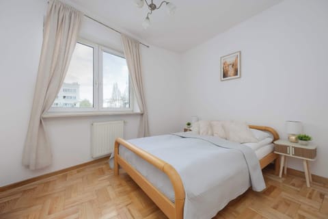 A bright bedroom with a wooden bed, white bedding, sheer curtains, and a small nightstand by the window.